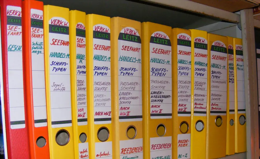 Reihe bunter Aktenordner mit beschrifteten Rückenschildern in einem Regal
