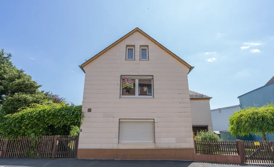 Giebelseite eines älteren Einfamilienhauses mit hell verkleideter Fassade und Rollladen, im Fenster ein Verkaufsschild, davor ein Holzzaun