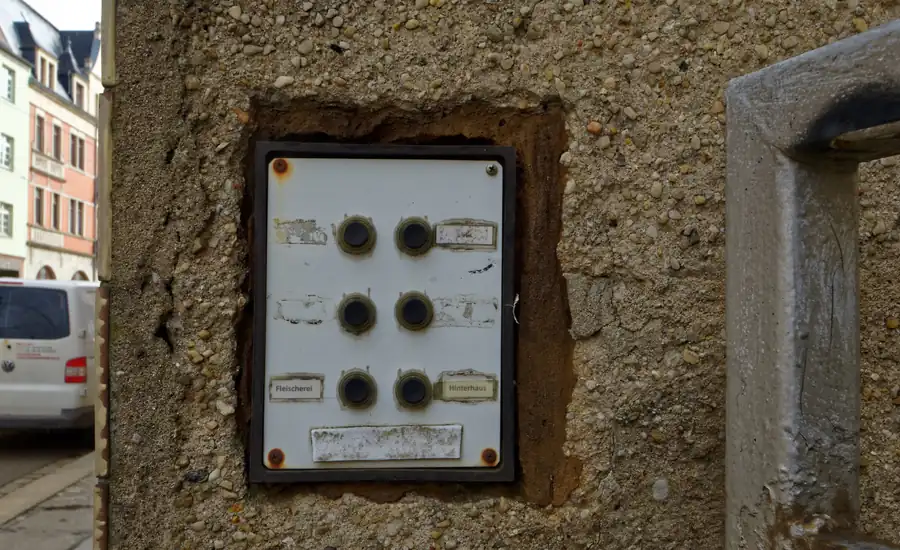 Verwitterte Klingeltafel mit sechs Klingelknöpfen und größtenteils leeren oder abgekratzten Namensschildern in der rauen Putzwand eines Altbau-Mehrfamilienhauses