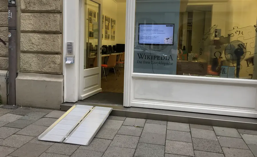 Offene Glastür eines Ladenlokals in einem Altbau, davor eine niedrige Eingangsstufe, über die eine zweiteilige Aluminium-Rampe auf den Gehweg führt