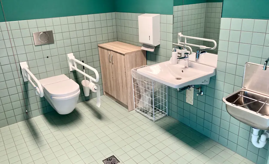 Barrierefreies WC mit Stützklappgriffen beidseits der wandhängenden Toilette, Waschbecken, hellgrün gefliesten Wänden und Bodenablauf