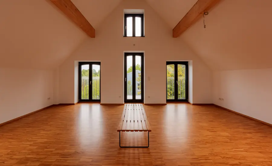Leere Dachgeschosswohnung mit glänzendem Parkett, weißen Wänden, sichtbaren Holzbalken und drei bodentiefen Fenstern, in der Mitte eine schmale Holzbank