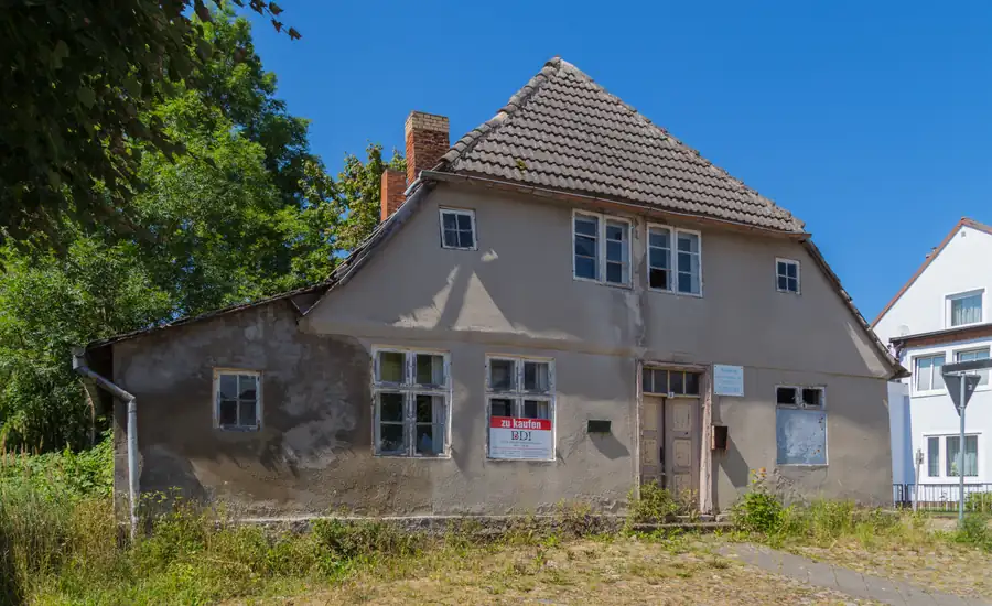 Leerstehendes älteres Wohnhaus mit grauem, stellenweise abgeplatztem Putz und Ziegeldach, im Fenster ein Schild „zu kaufen“, davor hohes Gras