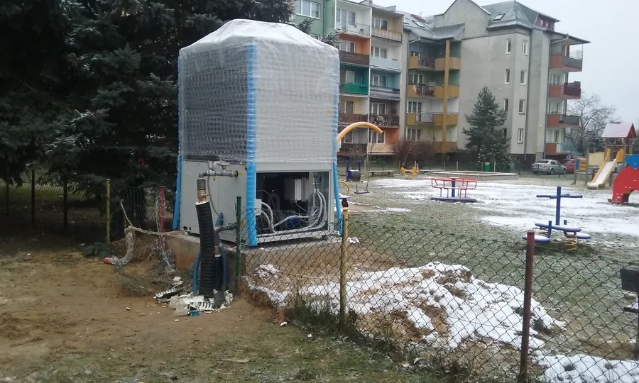 Luft-Wasser-Wärmepumpe mit Schutzfolie auf einem Betonsockel neben einem Spielplatz, dahinter ein mehrgeschossiges Wohnhaus mit bunten Balkonen