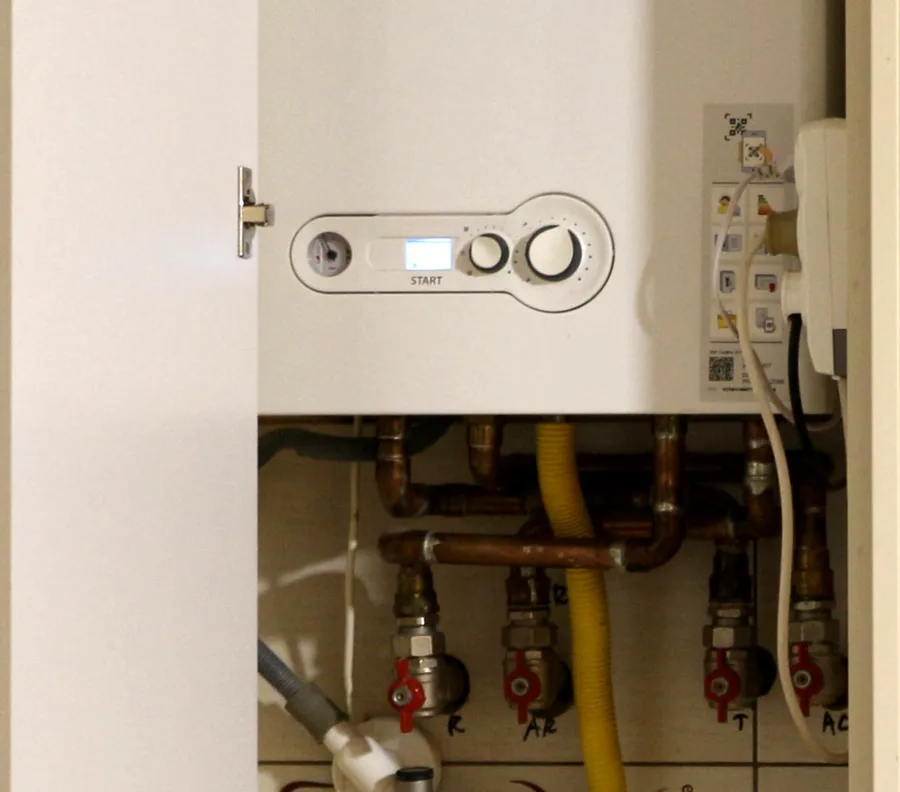 Wandhängende Gastherme mit Bedienelementen und angeschlossenen Leitungen in einem geöffneten Küchenschrank