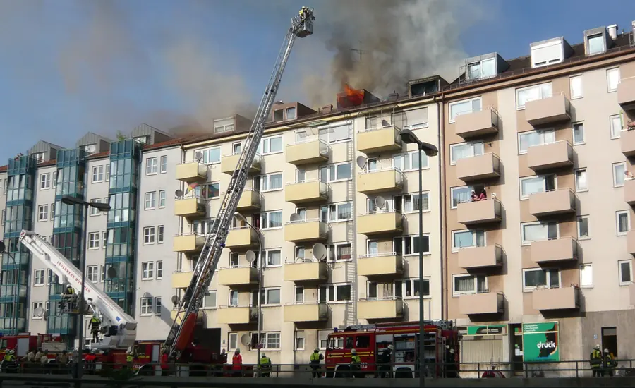 Brennendes Mehrfamilienhaus mit Rauch aus dem Dachgeschoss und ausgefahrener Drehleiter der Feuerwehr