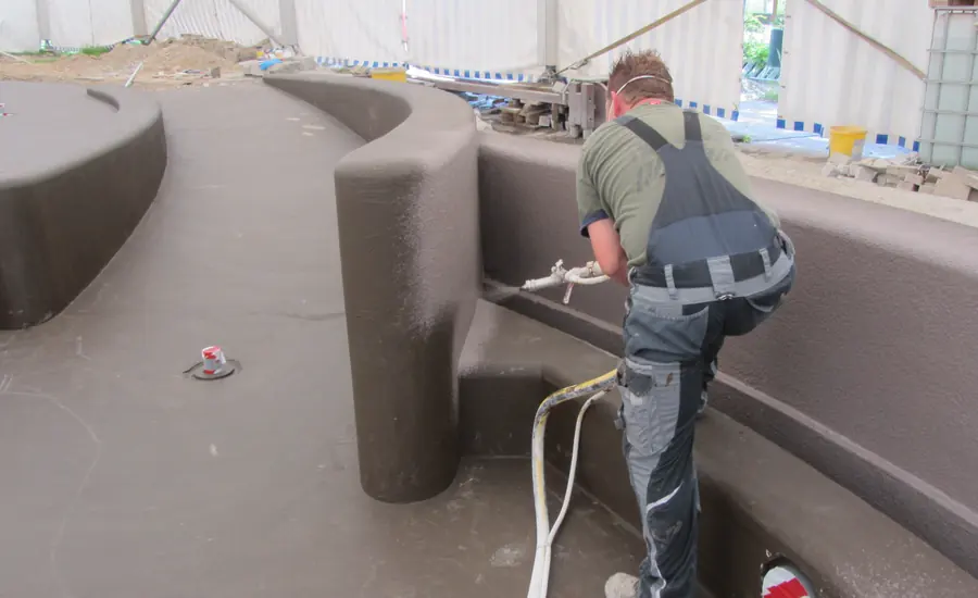 Arbeiter trägt mit einer Spritzpistole graue Dichtschlämme auf eine gerundete Betonwand auf
