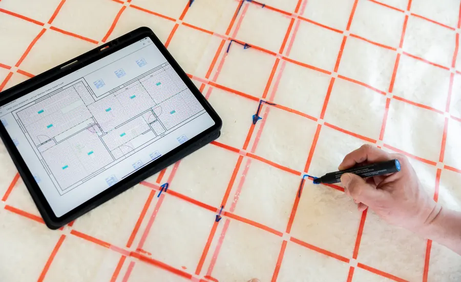 Tablet mit einem digitalen Grundriss liegt auf einer Rasterfolie für die Fußbodenheizung, daneben markiert eine Hand mit einem Stift eine Position