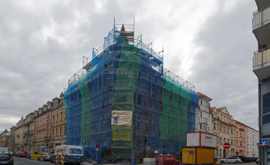 Mehrgeschossiges Eckhaus vollständig mit Gerüst und grünen Netzen eingehüllt, davor Baucontainer am Straßenrand