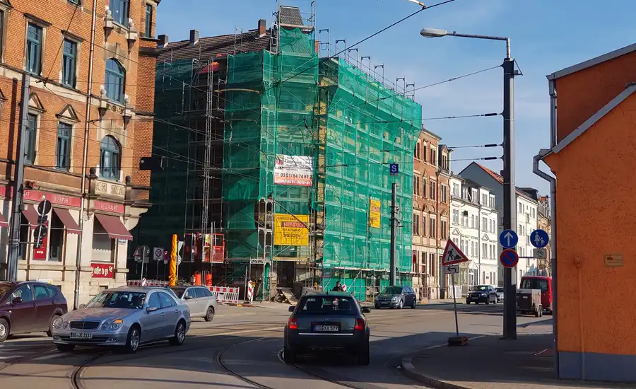 Eckhaus in der Sanierung, vollständig mit Baugerüst und grünem Gerüstnetz verhängt, davor eine Straße mit Autos