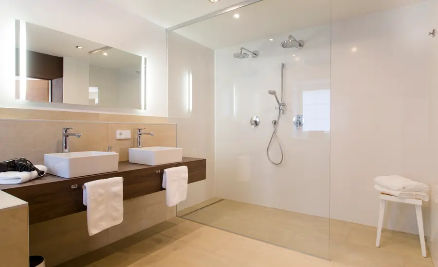 Modernes Badezimmer mit bodengleicher, verglaster Dusche, Doppelwaschtisch, Steckdose zwischen den Armaturen und beleuchtetem Spiegel
