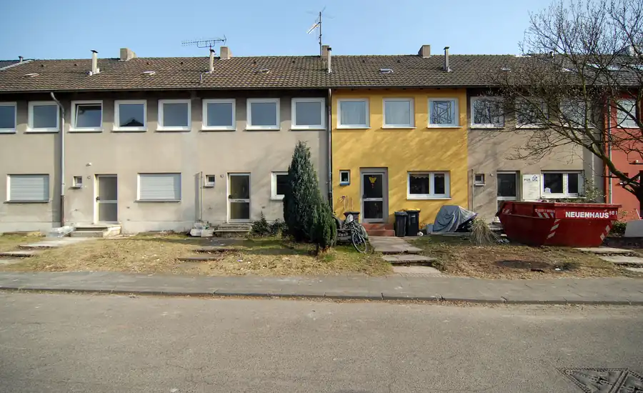 Reihenhauszeile aus der Nachkriegszeit mit grauen, gelben und roten Fassaden, davor schmale Vorgärten und ein Bauschuttcontainer