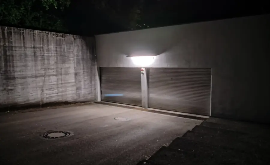 Garagenhof zwischen Betonwänden bei Nacht: zwei geschlossene Rolltore unter einer Außenleuchte, davor Schachtdeckel im Asphalt