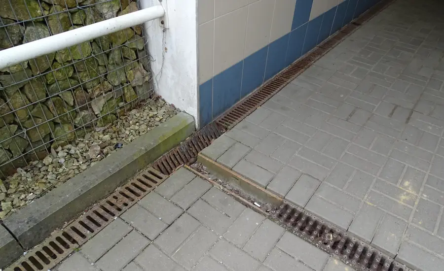 Entwässerungsrinnen mit rostigen Gitterrosten im Betonsteinpflaster: Eine verläuft direkt an einer gefliesten Wand entlang, eine zweite quer über den Weg
