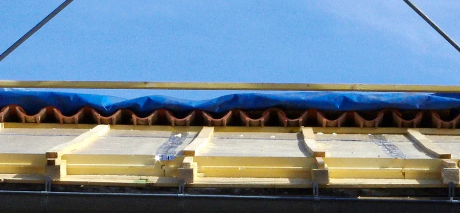 Dachdetail mit Dämmplatten und Holzlatten vor einer Reihe Dachziegel und einer blauen Plane