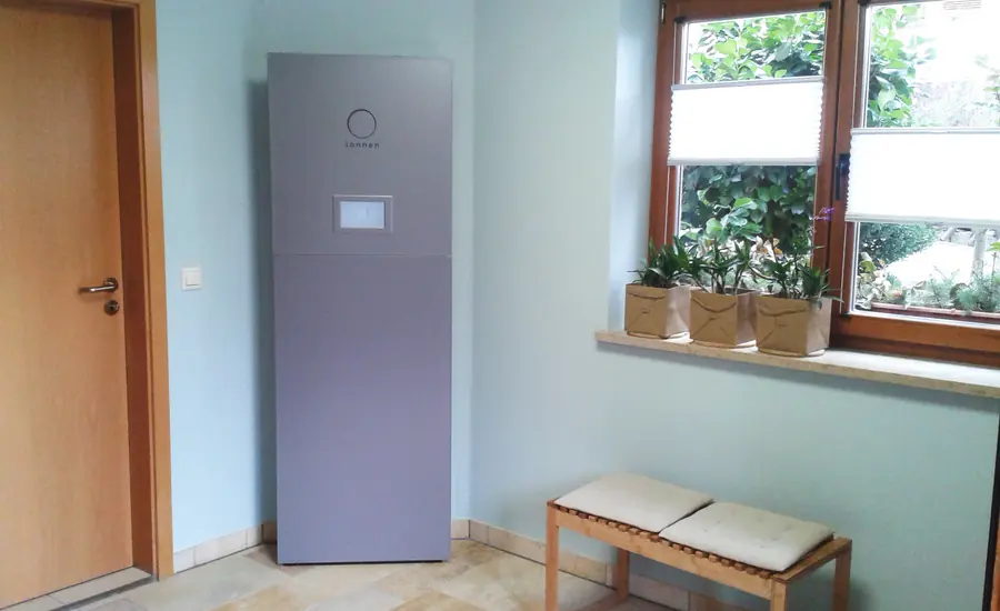 Grauer Batteriespeicher im Schrankformat mit kleinem Display, aufgestellt in einem hellen Raum eines Wohnhauses neben Tür und Fenster
