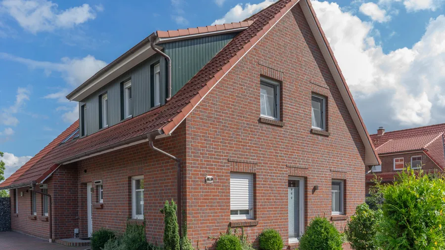 Freistehendes Backsteinhaus mit rotem Satteldach, grüner Gaube und Vorgarten unter blauem Himmel; Symbolbild für ein Ferienhaus