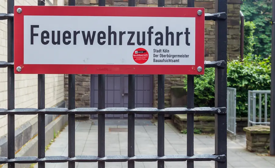 Rot-weißes Schild mit der Aufschrift Feuerwehrzufahrt an einem schwarzen Metallgitterzaun vor einer Einfahrt