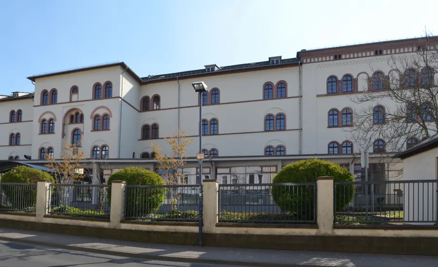 Langgestrecktes weißes Verwaltungsgebäude mit Rundbogenfenstern hinter einem Zaun mit Hecken