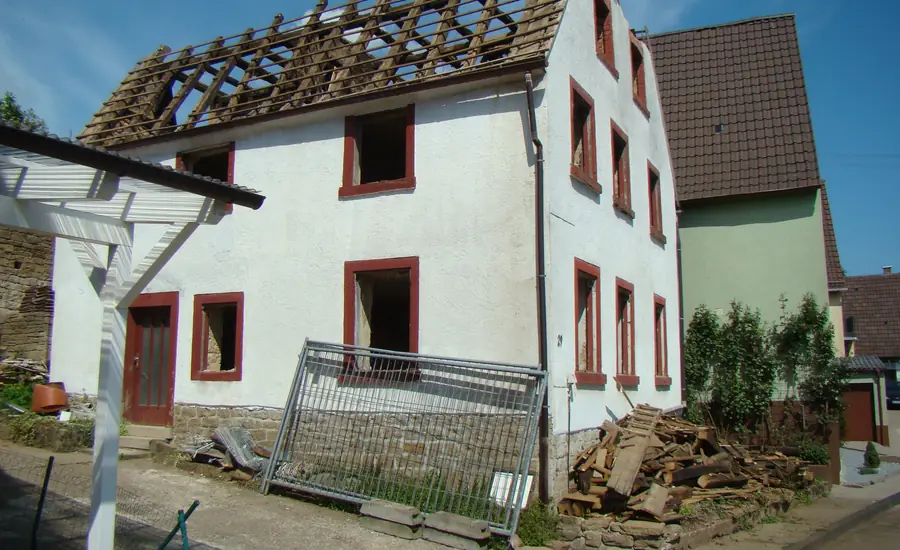 Altes Wohnhaus in der Kernsanierung: Dach bis auf die Sparren abgedeckt, Fenster ausgebaut, davor ein Bauzaunelement und ein Stapel alter Holzbalken