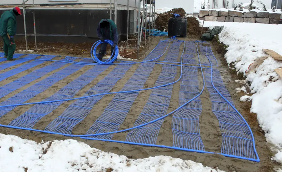 Blaue Kunststoffrohre eines Flächenkollektors, in dichten Schleifen auf Sandboden neben einem Neubau verlegt, am Rand Schnee, im Hintergrund zwei Arbeiter