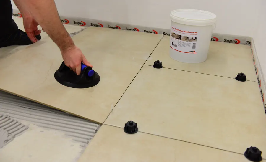 Hand drückt eine große Bodenfliese mit einem Saugheber ins Kleberbett, daneben schwarze Nivellierhilfen und ein Eimer