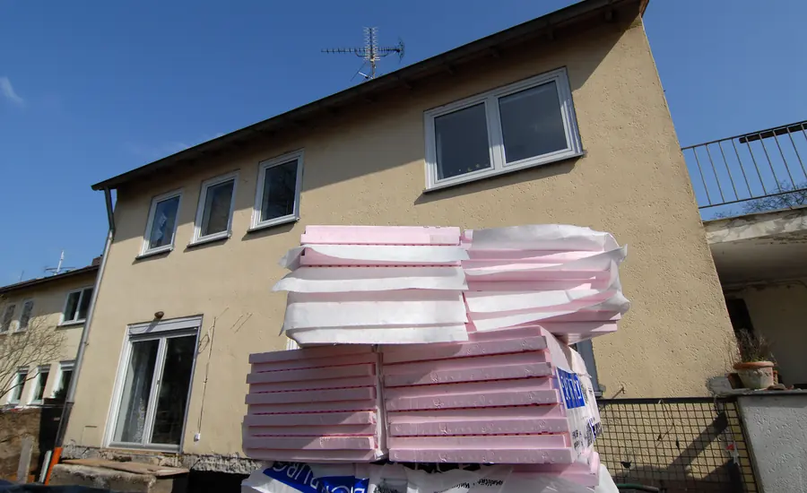 Zweigeschossiges Wohnhaus mit gelbem Altputz, davor auf Paletten gestapelte rosa Dämmplatten für die Fassade
