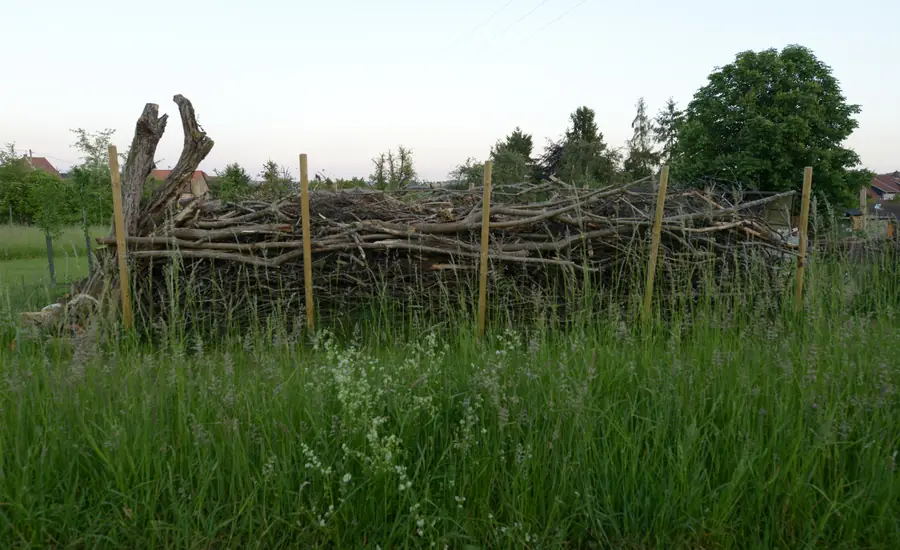Totholzhecke aus geschichteten Ästen zwischen zwei Reihen Holzpfosten auf einer Wiese