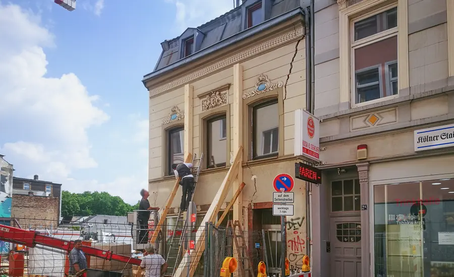 Wohn- und Geschäftshaus, das mit schrägen Holzbalken abgestützt wird, daneben eine offene Baustelle mit Bauarbeitern