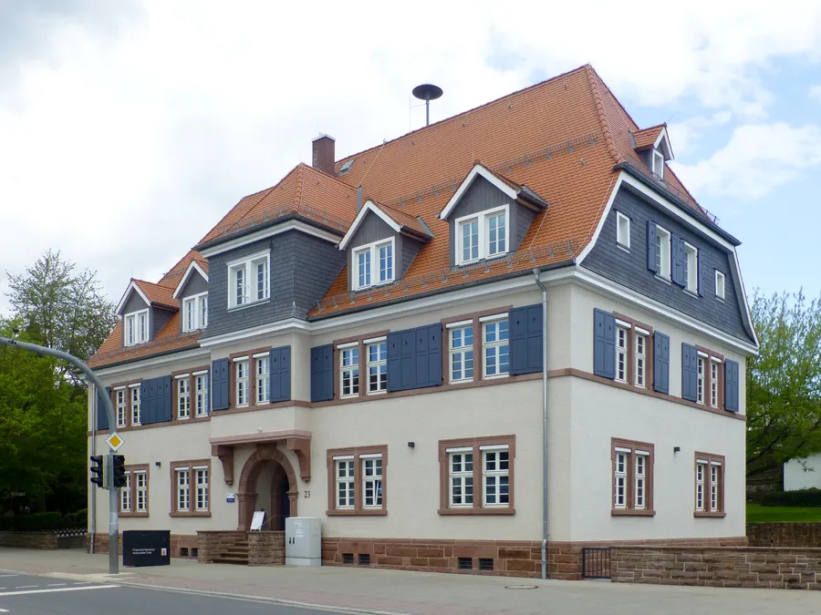 Zweigeschossiges Amtsgebäude mit hellem Putz, grauen Schieferflächen, roten Dachziegeln und blauen Fensterläden an einer Straßenecke