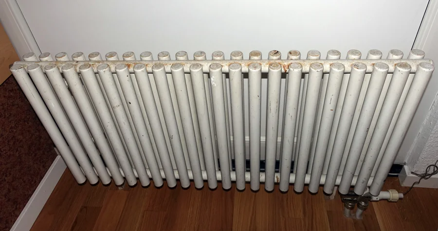 Weißer Röhrenheizkörper mit Gebrauchsspuren und Thermostatventil vor einer Wand.