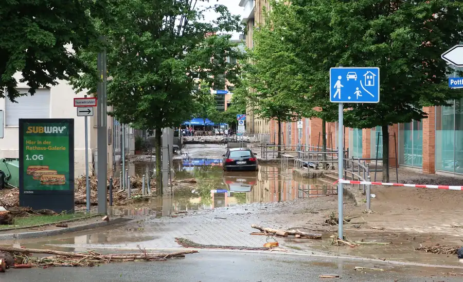 Überflutete Straße mit Schlamm, angeschwemmtem Treibgut und einem im Wasser stehenden Auto zwischen Häusern