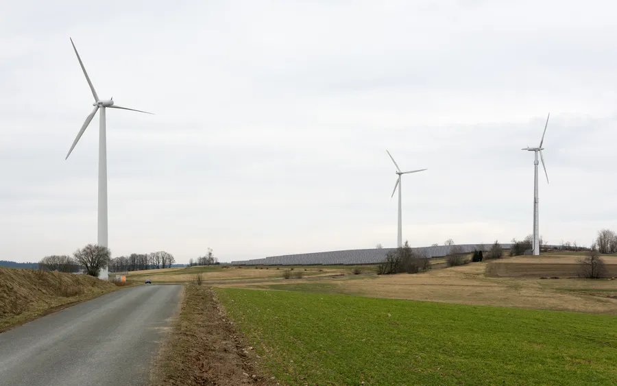 Drei Windräder neben einer Freiflächen-Solaranlage auf einer Hügelkuppe, davor ein Feldweg und Ackerland