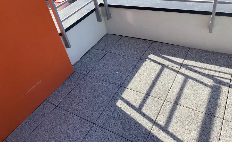 Gesprenkelte graue Terrassenplatten mit offenen Fugen auf einer Dachterrasse, dahinter die Attika mit Blechabdeckung und Geländer, links eine orangefarbene Wandplatte