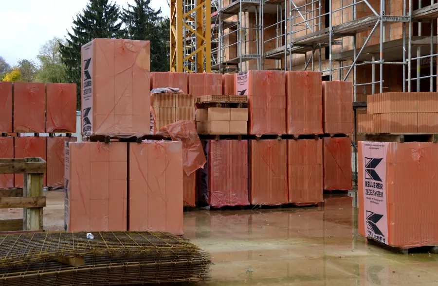 In rote Folie verpackte Paletten mit Hochlochziegeln auf einer nassen Baustellenfläche, dahinter ein eingerüsteter Mauerwerksrohbau und ein gelber Baukran