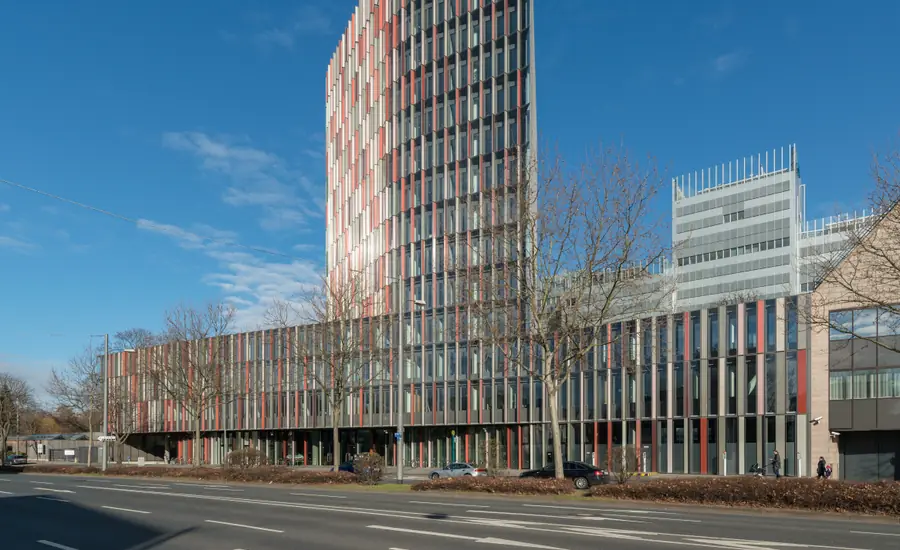 Bürogebäude mit farbiger Lamellenfassade und angeschlossenem Hochhaus an einer Straße mit kahlen Bäumen