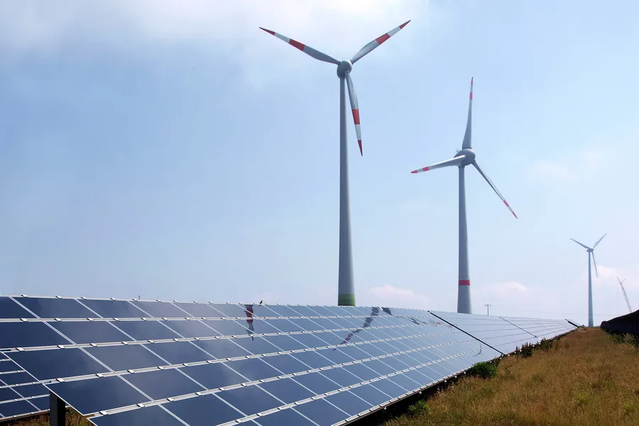 Reihen von Solarmodulen auf einer Wiese vor drei Windkraftanlagen unter leicht bewölktem Himmel
