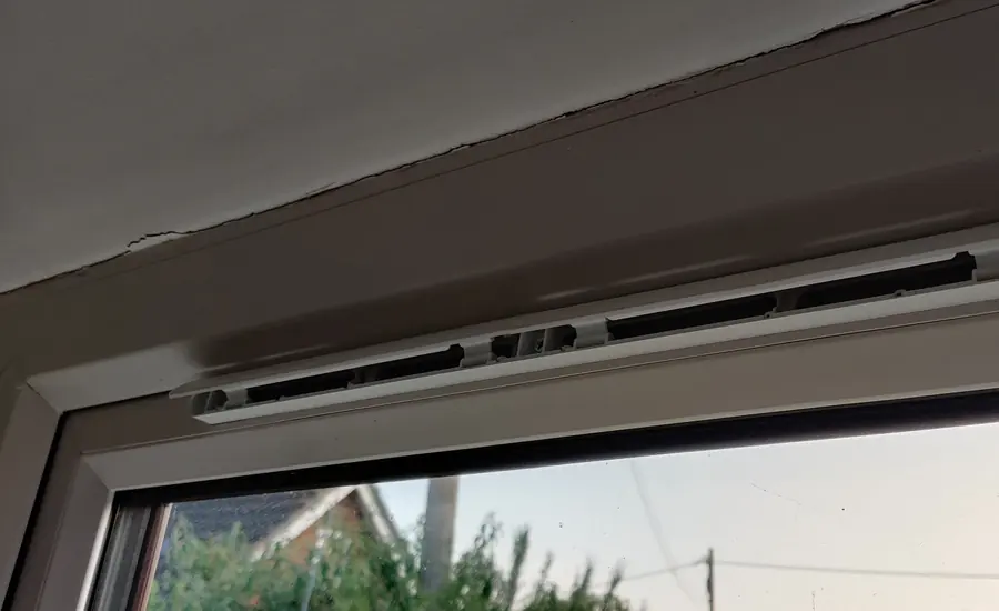 Geöffneter Fensterfalzlüfter oben im Rahmen eines weißen Kunststofffensters, dahinter Hausdächer