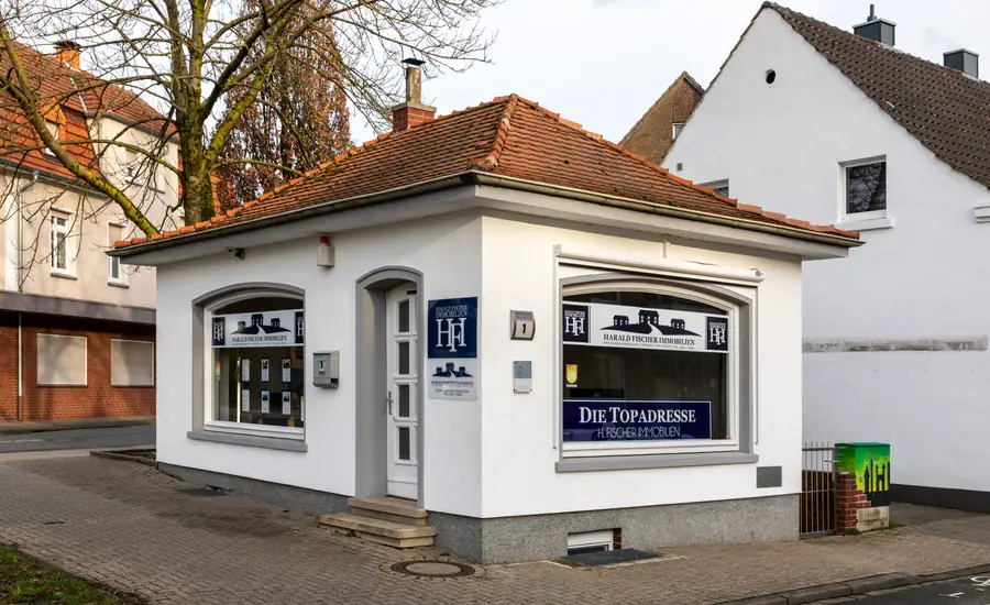 Kleines weißes Büro eines Immobilienmaklers mit Walmdach und beschrifteten Schaufenstern an einer Straßenecke in Dülmen