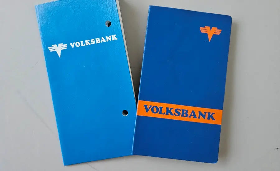 Zwei blaue Sparbücher der Volksbank auf grauem Untergrund, das rechte mit orangefarbenem Schriftband, das linke gelocht
