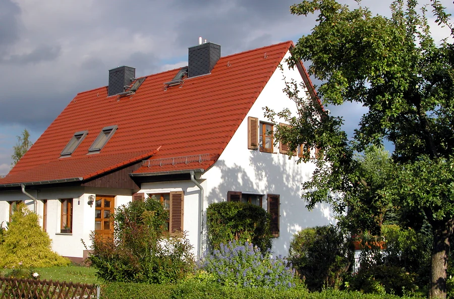 Weiß verputztes Haus mit rotem Ziegeldach, Dachfenstern und braunen Fensterläden hinter Sträuchern und Bäumen