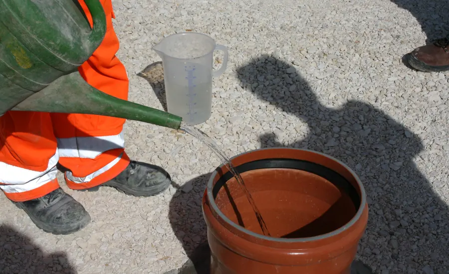 Arbeiter in orangefarbener Warnkleidung gießt mit einer Gießkanne Wasser in ein eingegrabenes Kunststoffrohr im Kiesboden, daneben ein Messbecher