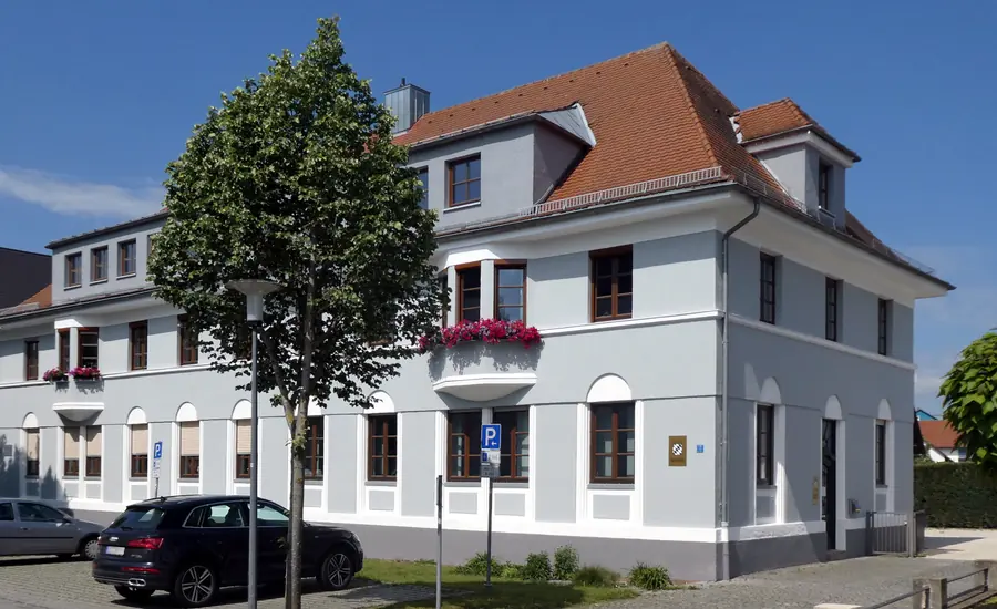 Hell verputztes Bürogebäude eines Notariats mit Walmdach und Blumenkästen, neben dem Eingang ein goldenes Schild mit bayerischem Rautenwappen und der Aufschrift Notar