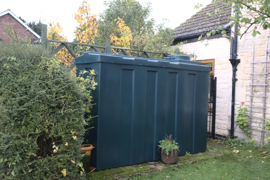 Dunkelgrüner Kunststofftank für Heizöl im Garten neben einem Wohnhaus, davor ein Blumentopf, links eine Hecke