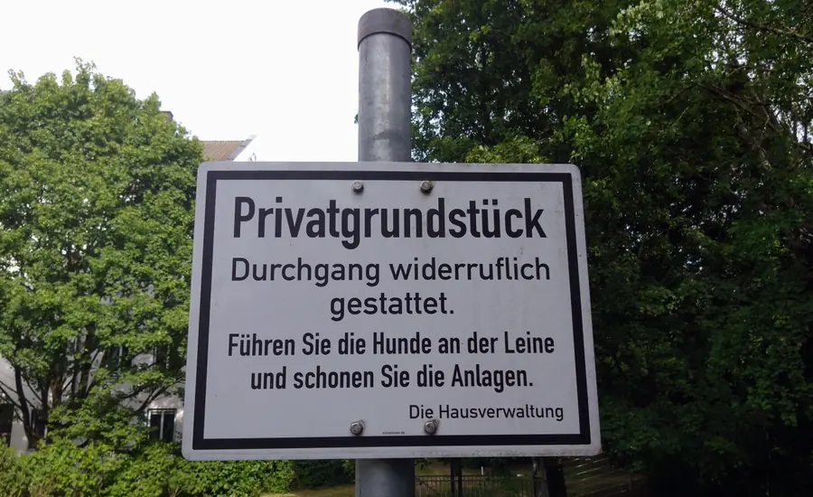 Schild an einem Pfosten mit der Aufschrift Privatgrundstück, Durchgang widerruflich gestattet, dahinter Hecke und Bäume