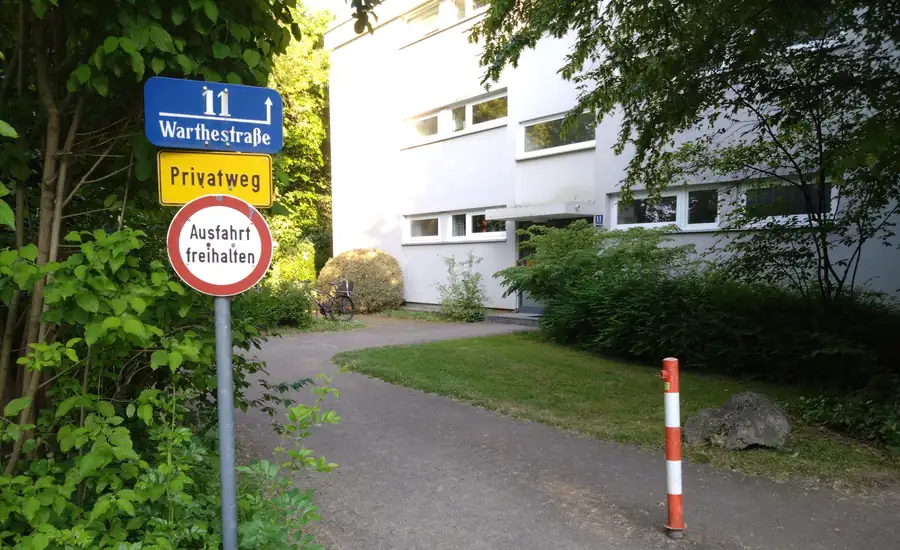 Gelbes Schild mit der Aufschrift Privatweg an einem schmalen asphaltierten Weg neben einem Wohnhaus