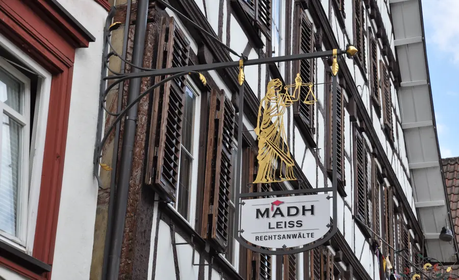 Schmiedeeiserner Ausleger mit vergoldeter Justitia-Figur und einem Schild mit der Aufschrift Rechtsanwälte an einer Fachwerkfassade