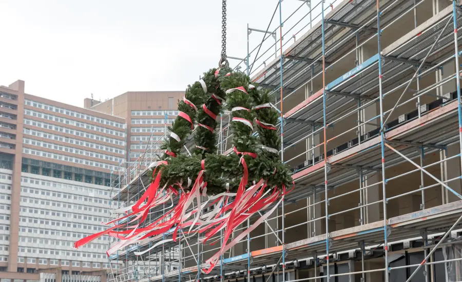 Mit roten und weißen Bändern geschmückte Richtkrone, die an einem Seil vor einem eingerüsteten Neubau hängt