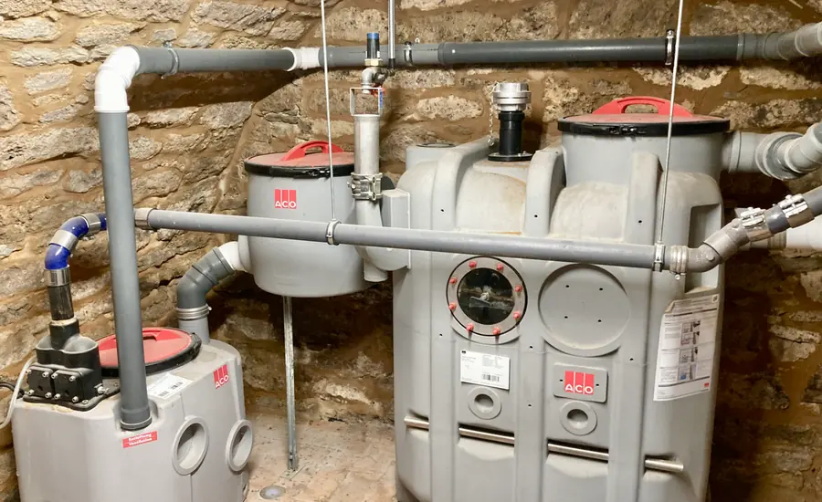 Graue Kunststoffbehälter einer Abwasserhebeanlage mit angeschlossenen Rohrleitungen vor einer Bruchsteinwand im Keller