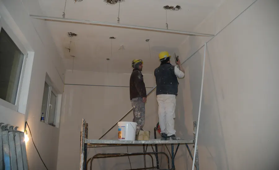 Zwei Bauarbeiter mit Schutzhelmen auf einem Rollgerüst bei Arbeiten an einer Innenwand